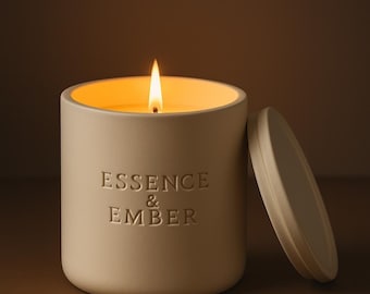May include: A lit, cream-colored candle in a ceramic container with the words "Essence & Ember" embossed on the side. The lid is off to the side. The candle's flame glows brightly against a warm background.