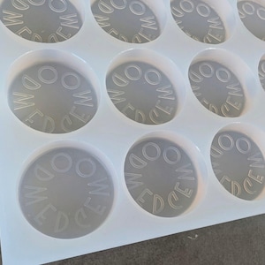 May include: A clear, flexible silicone mold with multiple circular cavities. Each cavity has the letters "WEM" and "EDO" in a circular pattern. The mold is designed for crafting or casting small, round objects.