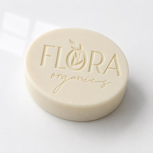 May include: An oval-shaped off-white bar of soap featuring the embossed text FLORA organics with a small leaf icon integrated into the letter O. The surface is smooth and matte, with the branding clearly stamped into the center of the bar.
