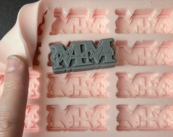 May include: A pink silicone mould with 12 impressions of the text "MM, Marie & Moods". The mould is partially open, revealing a grey silicone impression of the text.
