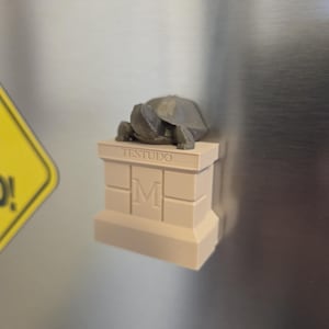 Peut inclure: Une petite figurine de tortue en 3D de couleur marron sur une base en 3D beige avec le mot "TESTUDO" et la lettre "M" dessus.