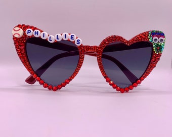 May include: Red heart-shaped sunglasses with rhinestones and the word "PHILLIES" spelled out in white letters with blue outlines. A baseball icon is on the left side of the sunglasses.