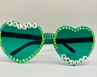 May include: Heart-shaped sunglasses with green lenses and a sparkly green frame. The frame is decorated with white beads that spell out "PHILLY" on both sides.