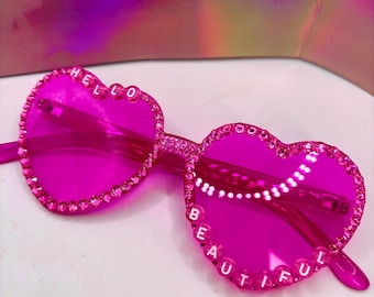 May include: Bright pink heart-shaped sunglasses with the words "HELLO BEAUTIFUL" spelled out in white letters. The frames are embellished with small, sparkling rhinestones. The lenses are a solid, vibrant pink.