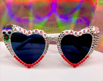 May include: White heart-shaped sunglasses with rhinestones and red beads. The sunglasses have the words "Atta Boy Harper" spelled out in white letters on the frames.