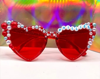 May include: Red heart-shaped sunglasses with white and blue pearl embellishments. The sunglasses have the word "PHILLIES" spelled out in white letters on the frame.