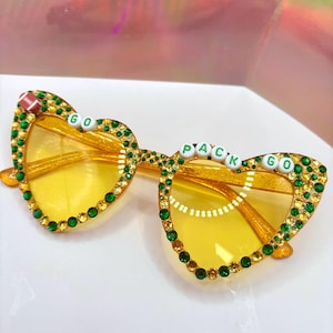 May include: Yellow heart-shaped sunglasses with green rhinestones and white letters spelling "GO PACK GO" on the frames.