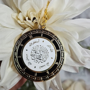 May include: A round, gold-toned pendant with black and white detailing, featuring Arabic calligraphy. The pendant is attached to a gold chain and is set against a backdrop of white flowers. The design includes a clock-like border.