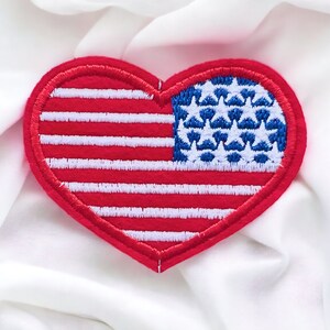 Könnte beinhalten: Ein herzförmiger Aufnäher mit dem Design der amerikanischen Flagge. Der Aufnäher hat einen roten Rand und rot-weiß gestreifte Muster. Die obere linke Ecke zeigt ein blaues Feld mit weißen Sternen.