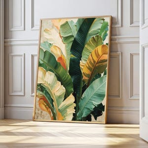 May include: A framed botanical art print depicting banana leaves. The artwork features a palette of greens, yellows, and oranges, with textured brushstrokes. The print is displayed against a white wall and wooden floor. The frame is made of wood.