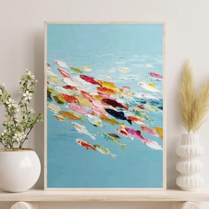 Op de afbeelding: Abstract schilderij van een school vissen in tinten rood, geel, roze en wit, zwemmend in een lichtblauwe zee. Het kunstwerk is ingelijst en wordt tentoongesteld op een witte plank met decoratieve elementen.