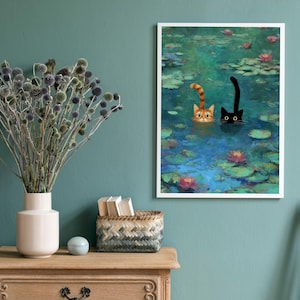 May include: A framed art print showing two cats in a pond with lily pads and flowers. One cat is ginger and white, the other is black. The artwork is displayed on a teal wall, with a wooden table and vase of dried flowers in the foreground.