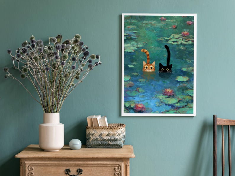 May include: A framed art print showing two cats in a pond with lily pads and flowers. One cat is ginger and white, the other is black. The artwork is displayed on a teal wall, with a wooden table and vase of dried flowers in the foreground.