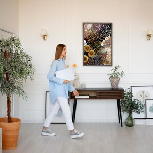Puede incluir: Una persona con una camisa azul claro y pantalones blancos camina por una habitaci&oacute;n luminosa, sosteniendo una computadora port&aacute;til blanca. Una impresi&oacute;n floral enmarcada cuelga de la pared sobre una consola de madera oscura. Plantas decorativas y obras de arte completan el interior.