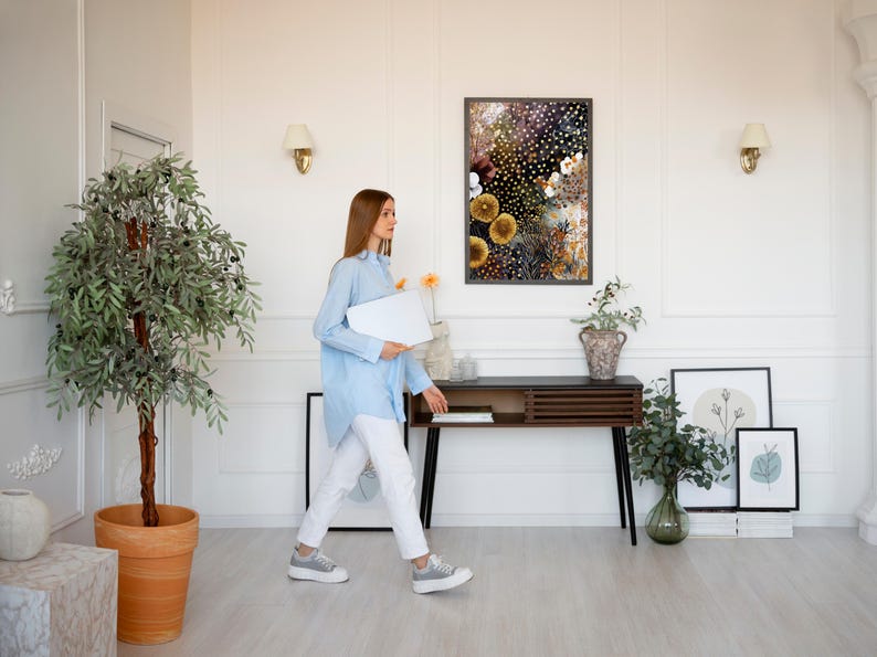 Puede incluir: Una persona con una camisa azul claro y pantalones blancos camina por una habitaci&oacute;n luminosa, sosteniendo una computadora port&aacute;til blanca. Una impresi&oacute;n floral enmarcada cuelga de la pared sobre una consola de madera oscura. Plantas decorativas y obras de arte completan el interior.