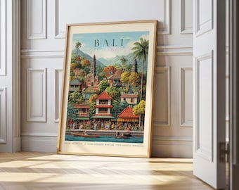 May include: A framed vintage travel poster of Bali, Indonesia, featuring a colorful illustration of a village with temples, palm trees, and a river. The poster has a light wood frame and is leaning against a white wall.