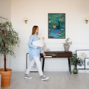 May include: A bright living room with a painting of cats in a pond, a wooden console table, and a large potted tree. The room has white walls, a light wood floor, and wall sconces. A person is walking through the room.