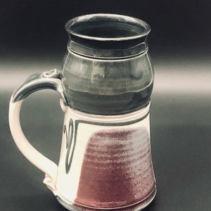 May include: A handmade ceramic mug with a unique design. The mug features a black top section, a white and burgundy lower section, and a white handle. The mug's design includes a black, stylized heart shape. The mug is a unique piece of pottery.
