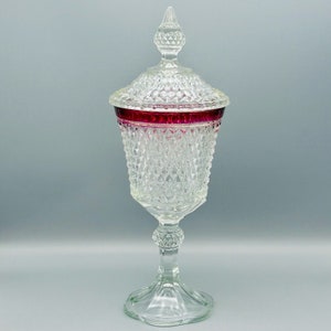May include: A clear, vintage glass candy dish with a diamond pattern and a ruby red rim. The dish has a lid with a pointed finial and sits on a pedestal base. A decorative piece for the home.