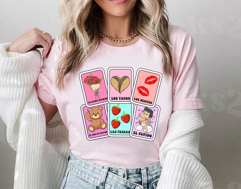May include: Pink t-shirt featuring a graphic of a Loteria card set. The cards depict images of a bouquet of flowers, tacos, lips, a teddy bear, strawberries, and a cupid. The text on the cards reads "El ramo de flores", "Los tacos", "Los besitos", "El oso de peluche", "Las fresas", and "El cupido".