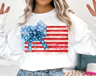 May include: White sweatshirt with a watercolor design of an American flag with a blue bow on top. The flag is red, white, and blue.
