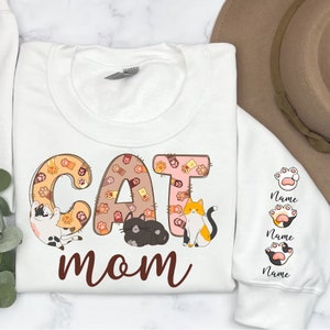 May include: White sweatshirt with the words "CAT MOM" in brown lettering. The word "CAT" is made up of paw print designs with a cat in each letter. There are three paw print designs on the sleeve with the word "Name" below each.