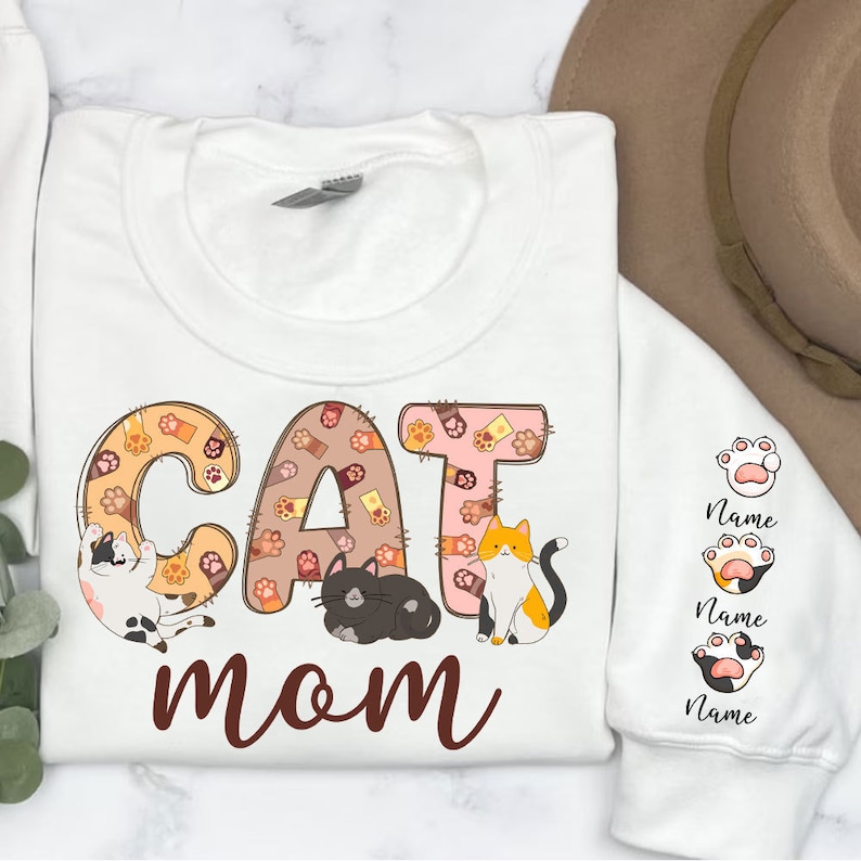 May include: White sweatshirt with the words "CAT MOM" in brown lettering. The word "CAT" is made up of paw print designs with a cat in each letter. There are three paw print designs on the sleeve with the word "Name" below each.