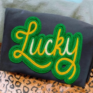May include: A dark gray sweatshirt with a large, embroidered "Lucky" patch. The patch features green, yellow, and white rope-like stitching, creating a textured effect. The word "Lucky" is in a playful, cursive font.