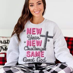 May include: A light gray sweatshirt with the text "NEW Year NEW Challenges SAME Great God" in pink and gray. A gray cross graphic is to the right of the text. The sweatshirt is a crew neck style.