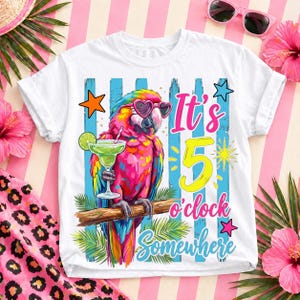May include: A white t-shirt featuring a colorful parrot wearing heart-shaped sunglasses and holding a lime-garnished cocktail. The graphic includes blue vertical stripes, palm leaves, stars, and the text It's 5 o'clock Somewhere in pink and yellow.