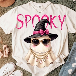 May include: A white short-sleeve t-shirt featuring the pink text SPOOKY above a graphic of a ghost wearing a black witch hat with a pink polka-dot bow, leopard-print sunglasses, and layered gold necklaces with white pearls and pink teardrop gems.
