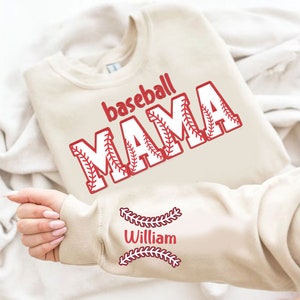 Peut inclure: Un sweat-shirt beige avec le texte "baseball MAMA" en rouge et blanc avec un motif de couture de baseball. La manche a le texte "William" en rouge avec un motif de couture de baseball.