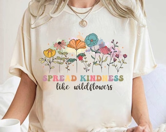 May include: Cream-coloured t-shirt with a colourful floral design and the text "SPREAD KINDNESS like wildflowers." The graphic features flowers in yellow, orange, blue, and pink. The shirt has a relaxed fit.