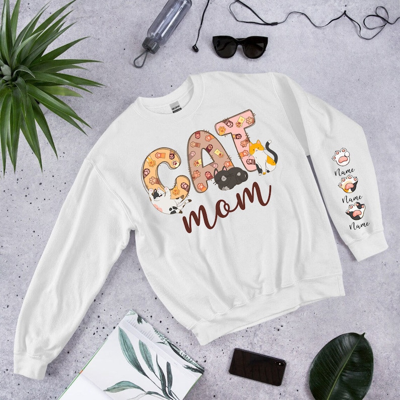 May include: White sweatshirt with the text "CAT MOM" in brown letters. The letters are decorated with images of cats. The sleeves have paw prints and the text "Name" repeated three times.
