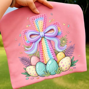 May include: A pink pillow with a colorful crochet cross design. The cross features a rainbow pattern and a purple bow. Easter eggs in various colors and patterns are at the base of the cross, with green foliage accents.
