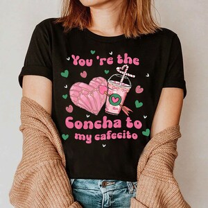May include: Black t-shirt with a pink heart-shaped pastry and a pink and green cup with a straw. The text on the shirt reads "You're the Concha to my cafecito".