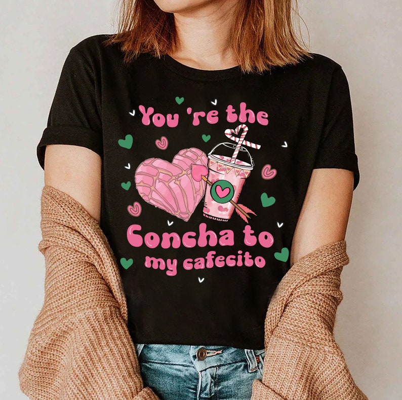 May include: Black t-shirt with a pink heart-shaped pastry and a pink and green cup with a straw. The text on the shirt reads "You're the Concha to my cafecito".