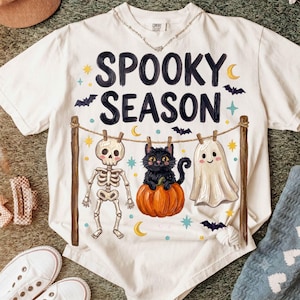 May include: A white short-sleeve t-shirt featuring the black text SPOOKY SEASON above a graphic of a clothesline holding a skeleton, a black cat sitting on an orange pumpkin, and a white ghost, accented with yellow stars, moons, and bats.