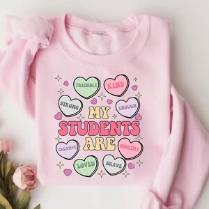 May include: Pink sweatshirt with a graphic design of hearts that say "My Students Are" with words like "Friendly", "Kind", "Strong", "Enough", "Creative", "Loved", "Brave", and "Worthy" inside each heart.
