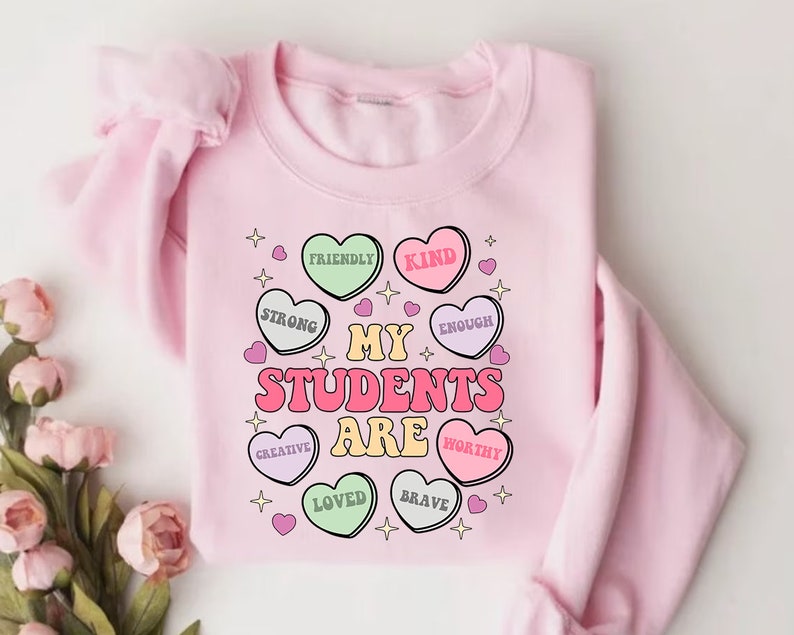 May include: Pink sweatshirt with a graphic design of hearts that say "My Students Are" with words like "Friendly", "Kind", "Strong", "Enough", "Creative", "Loved", "Brave", and "Worthy" inside each heart.