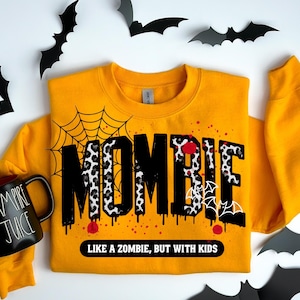 Puede incluir: Sudadera amarilla mostaza con la palabra "MOMBIE" en letras negras, estampadas de leopardo y blancas. La sudadera tiene un diseño de telaraña, salpicaduras rojas y la frase "LIKE A ZOMBIE, BUT WITH KIDS". Una taza negra con "VAMPIRE JUICE" está a la izquierda.