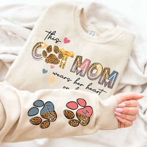 May include: Beige sweatshirt with the text "This Cat Mom wears her heart on her paw" with a paw print and a cat graphic. The sleeves have paw prints with the names "Hunter" and "Emily" on them.