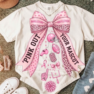 May include: A cream-colored short-sleeve t-shirt featuring a large pink bow graphic with leopard print accents. The text 'PINK OUT' and 'YOUR MASCOT' is printed on the bow tails. Below the bow are pink football-themed icons and charms.