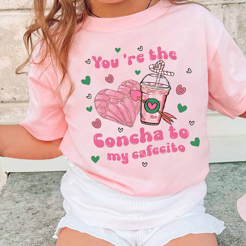 May include: Pink t-shirt with a graphic of a concha pastry and a cup of coffee with the text "You're the Concha to my cafecito".