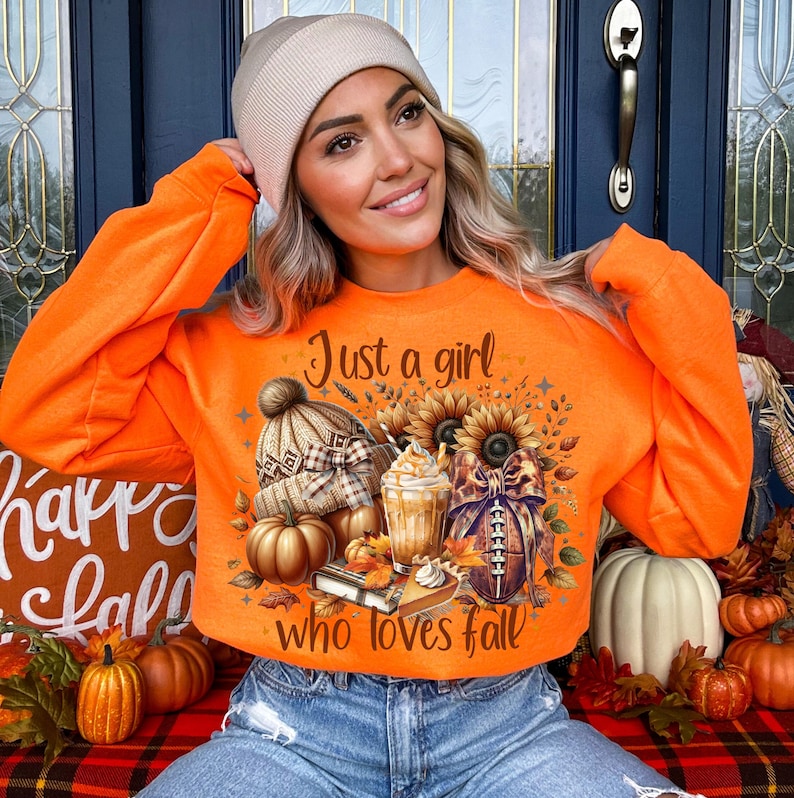 Pu&ograve; includere: Felpa arancione con il testo "Just a girl who loves fall" e una grafica di una zucca, una tazza di caff&egrave;, un pallone da football e un girasole.