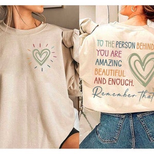 Puede incluir: Sudadera beige con un diseño de corazón colorido en la parte delantera y el texto "To the person behind me you are amazing beautiful and enough. Remember that" en la parte posterior.