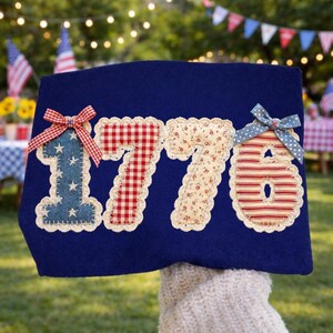 May include: A navy blue fabric decoration with the number "1776" appliqued in various patriotic patterns. The "1" is blue denim with white stars, the "7"s are red gingham and floral, and the "6" is red and white striped. Each number has a decorative bow.