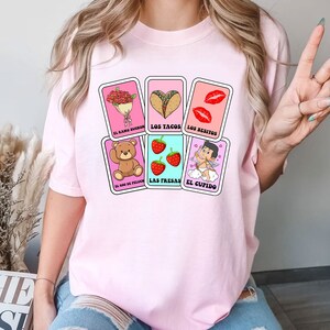 May include: Pink t-shirt with a graphic of five playing cards featuring illustrations of a bouquet of flowers, tacos, lips, strawberries, and a cupid. The cards have Spanish text on them: "El ramo de flores", "Los tacos", "Los besitos", "Las fresas", and "El cupido".