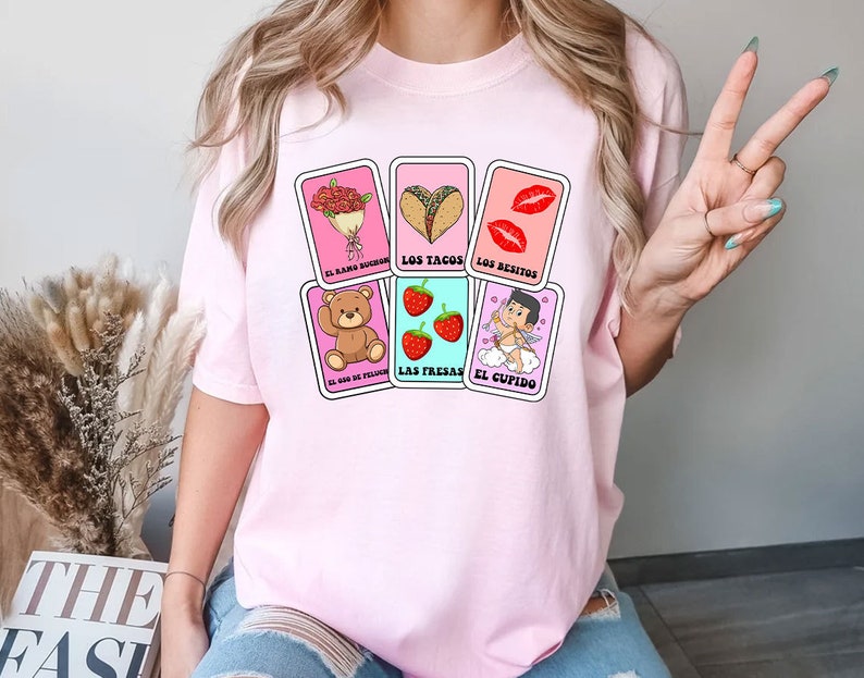 May include: Pink t-shirt with a graphic of five playing cards featuring illustrations of a bouquet of flowers, tacos, lips, strawberries, and a cupid. The cards have Spanish text on them: "El ramo de flores", "Los tacos", "Los besitos", "Las fresas", and "El cupido".