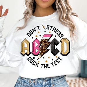 May include: White sweatshirt with a graphic print of a lightning bolt with a pencil inside, surrounded by stars and the text "Don't Stress ABC Rock the Test".